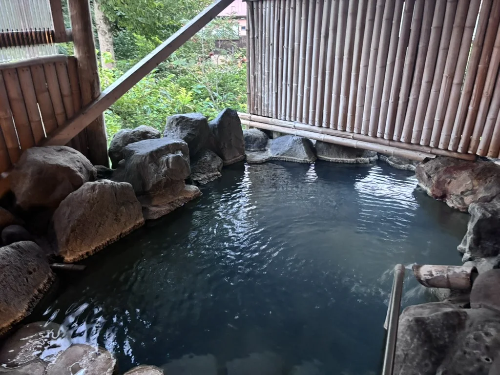 An outdoor rock bath at an Akakura inn at dusk, enclosed by a bamboo screen, a pipe feeding fresh hot spring water at the right