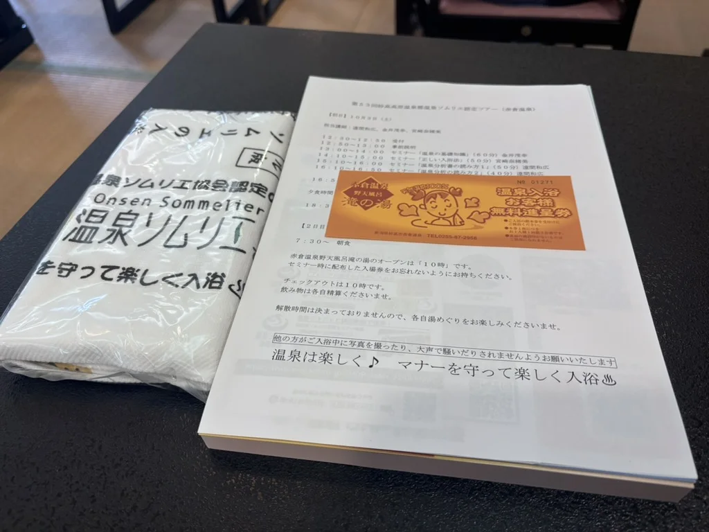 The day's timetable on a black desk, with a free-entry ticket for Akakura's Taki-no-yu open-air bath, next to the course towel still in its wrapper