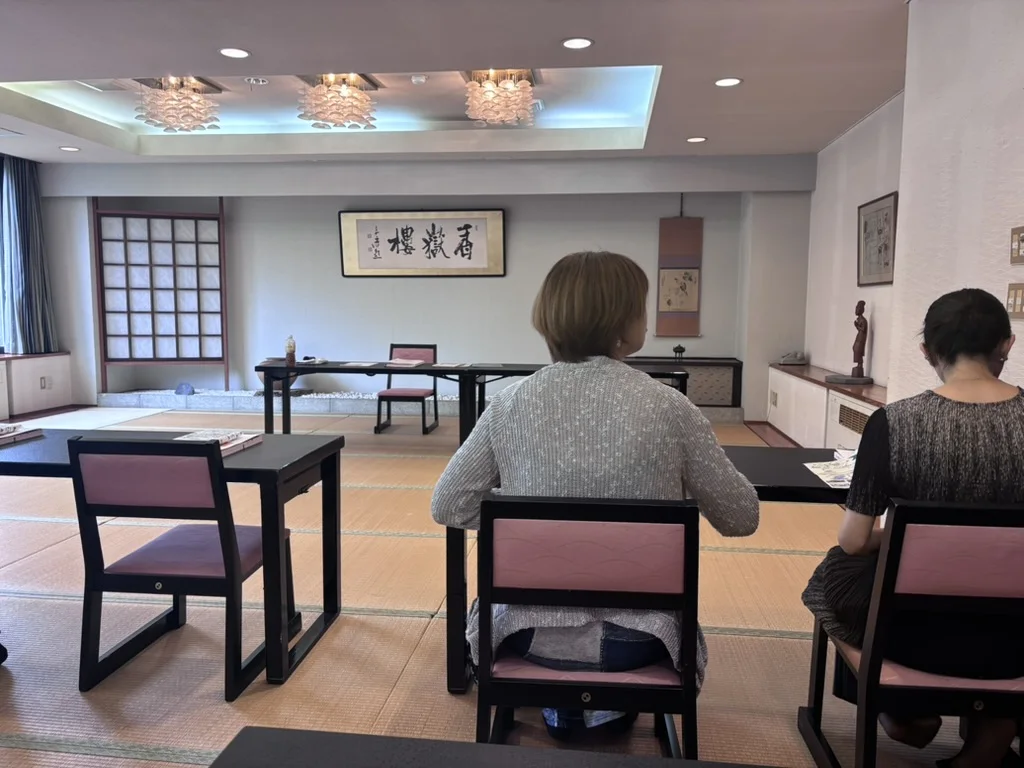A tatami-floored seminar hall in an Akakura inn, low black desks with pink chairs, a calligraphy scroll on the front wall and two participants waiting