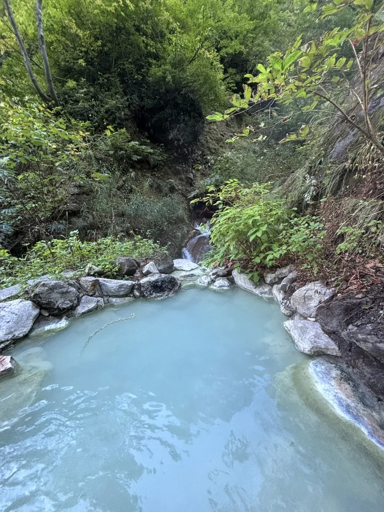A milky blue outdoor hot spring pool in a forested mountain gorge at Tsubame Onsen, a small stream running in behind it