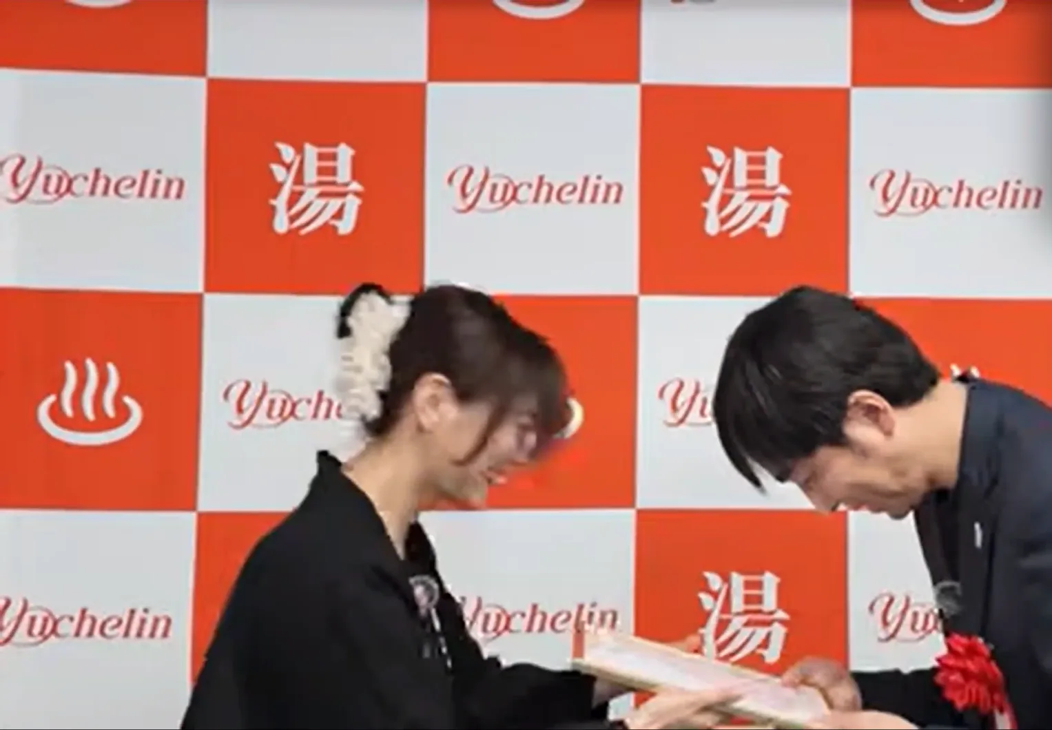 A presenter in black hands a certificate to a facility representative wearing a red rosette, in front of a backdrop printed with the Yuchelin logo and the character for hot water